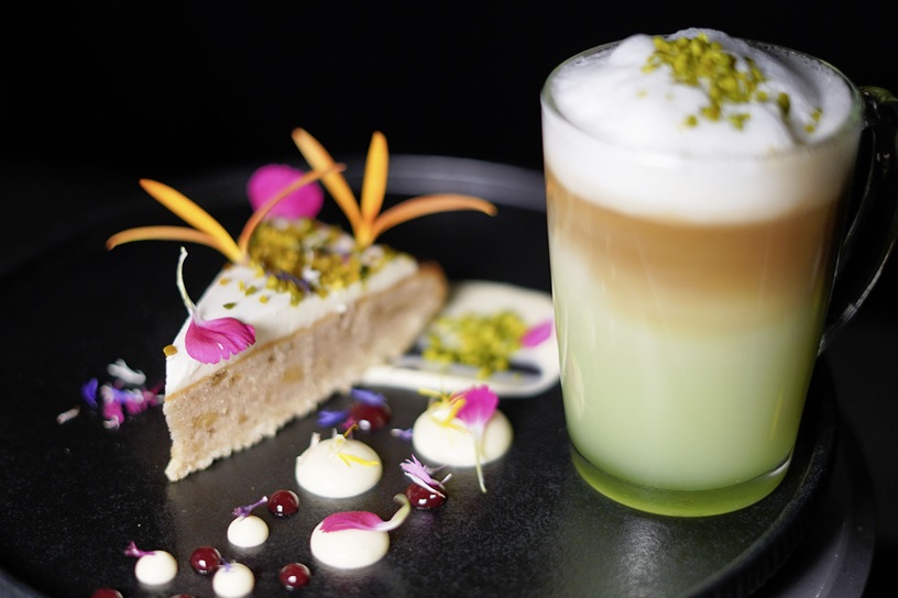 Banana Walnut Cake with pistachios and fresh edible petals, paired with a trendy latte